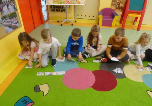 Sześcioro dzieci siedzi na dywanie z rozkłada przed sobą obrazkami, którym się przygląda.